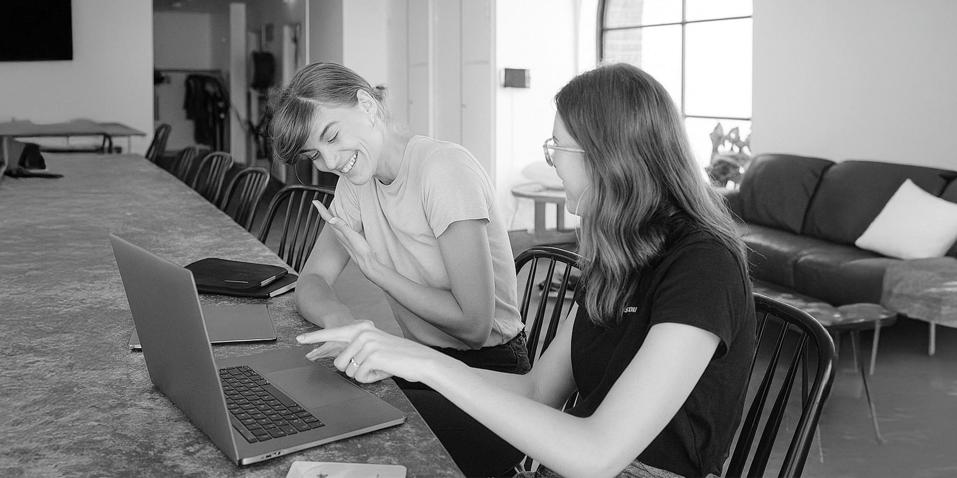 Dwie młode kobiety siedzą przy stole i pracują wspólnie na laptopie.