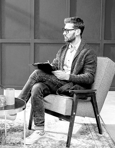 Mężczyzna w okularach siedzi w fotelu z notatnikiem na kolanach.